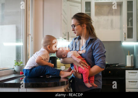 Young mother holds her little daughter with pacifier. Eldest son is sitting barefoot on table in kitchen and holding electronic thermometer. He kisses Stock Photo