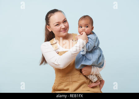 Portrait of a beautiful young mother in beige overalls holding adorable little baby girl in jeans dress in her arms against pale blue background. Baby Stock Photo