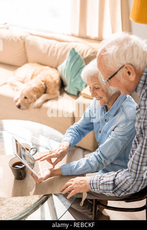 High angle  portrait of modern senior couple using digital tablet sitting at table at home with pet dog in background Stock Photo