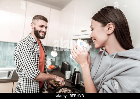 Smiling woman drinking tea while watching her man cooking Stock Photo