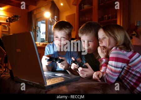 Siblings, 7, 11, 13 years old, with laptop computer in the living room, playing a car racing computer game Stock Photo