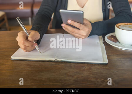 Young Asian lady using smartphone and writing notes at cafe Stock Photo