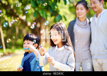 little asian kids boy and girl sister and brother blowing bubbles in a park with parents watching from behind. Stock Photo