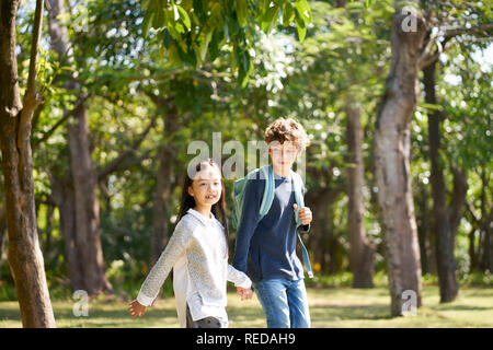 little asian girl and caucasian boy walking together holding hands outdoors in a park. Stock Photo
