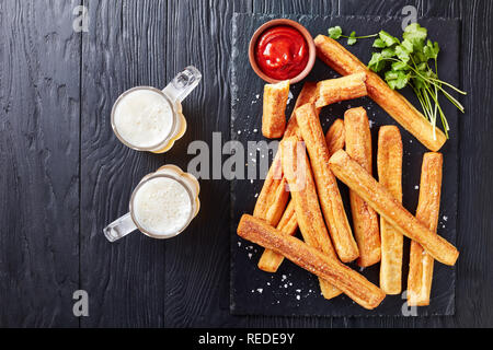 close-up of Cheese Straws, Cheesy Bread Sticks on a cutting wooden board with tomato sauce and two glass cups with foamed beer on a black wooden table Stock Photo