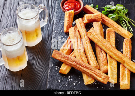 close-up of Cheese Straws, Cheesy Bread Sticks on a cutting wooden board with tomato sauce and two glass cups with foamed beer on a black wooden table Stock Photo