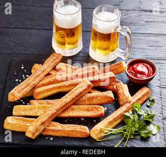 close-up of Cheese Straws, Cheesy Bread Sticks on a cutting wooden board with tomato sauce and two glass cups with foamed cold beer on a black wooden  Stock Photo