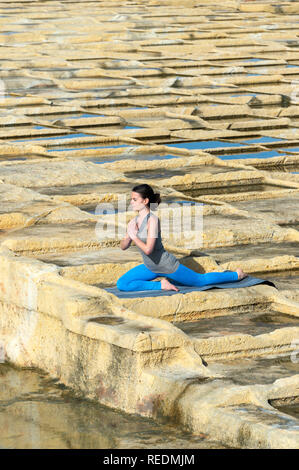 woman practicing yoga outside in the sunshine on the limestone salt pans, Malta, doing the pigeon pose. Stock Photo