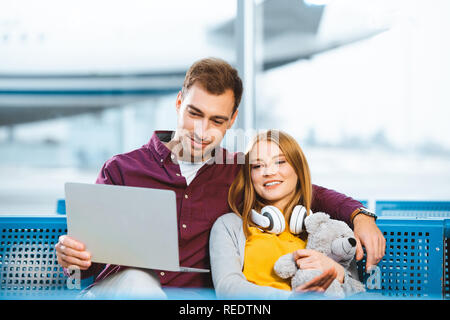 happy boyfriend using laptop near girlfriend with teddy bear in airport Stock Photo