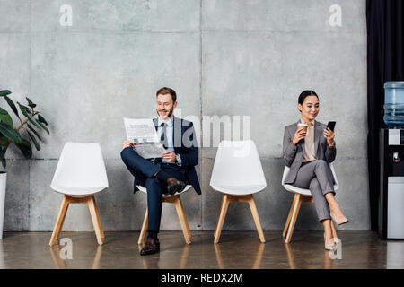 businessman reading newspaper while asian businesswoman using smartphone in waiting hall Stock Photo