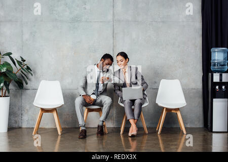 multiethnic colleagues with laptop and coffee sitting and checking time on watch in waiting hall Stock Photo