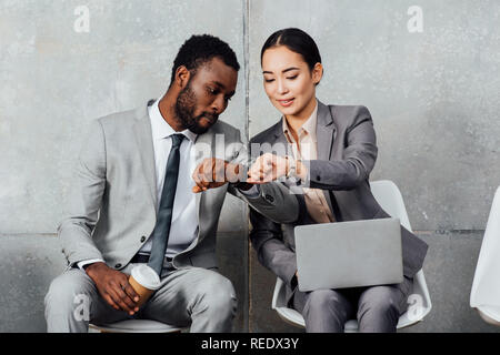 multiethnic colleagues with laptop and coffee sitting and checking time on watches in waiting hall Stock Photo