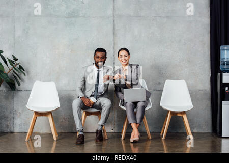 smiling multiethnic colleagues with laptop and coffee sitting and checking time on watches in waiting hall Stock Photo