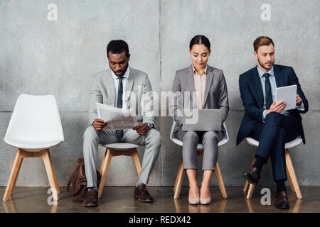 focused multiethnic businesspeople reading newspaper and using digital devices in waiting hall Stock Photo