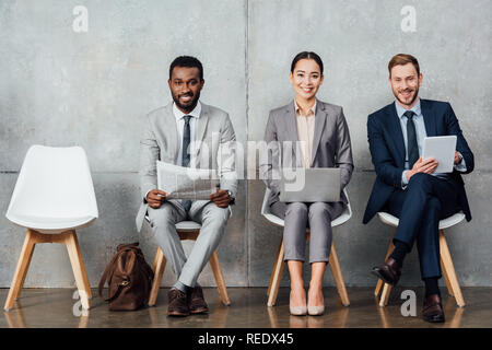 smiling multiethnic businesspeople reading newspaper and using digital devices in waiting hall Stock Photo