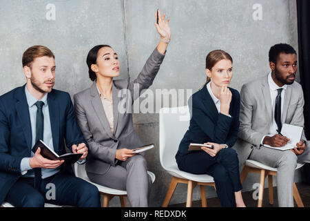 businesswoman raising hand while focused multiethnic businesspeople sitting on chairs with notebooks Stock Photo