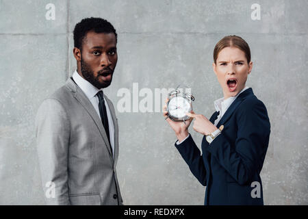 shocked businesswoman pointing at clock while african american businessman looking at camera on grey background Stock Photo