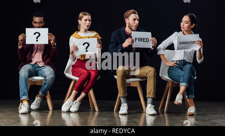 multiethnic people sitting on chairs and holding cards with 'director', 'fired' words and question marks isolated on black Stock Photo