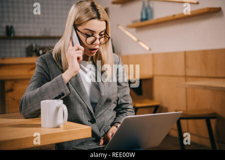 selective focus of shocked woman in glasses talking on smartphone while looking at laptop Stock Photo