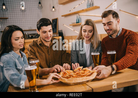 group of friends smiling while taking pieces of tasty pizza in bar Stock Photo