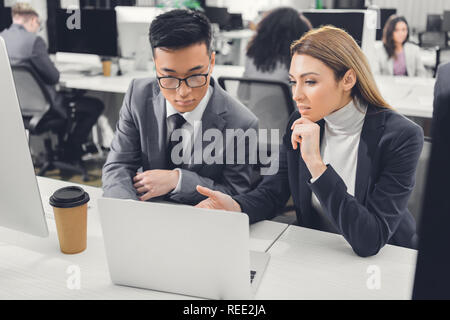 concentrated multiethnic businessman and businesswoman discussing work and pointing at laptop in office Stock Photo