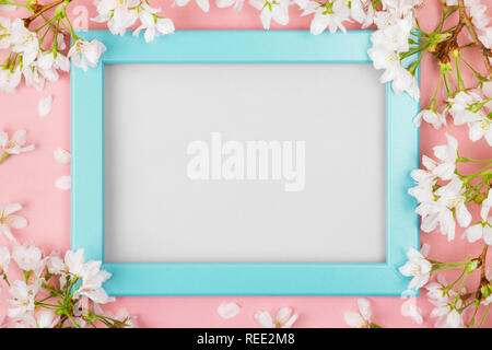Spring background with an empty turquoise picture frame surrounded by white cherry blossom flowers and branches on a pink surface. Flat lay Stock Photo