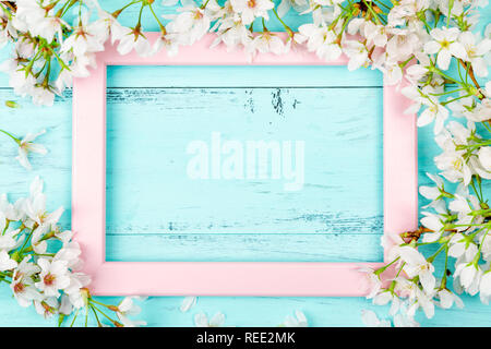 Spring background with an empty pink picture frame surrounded by white cherry blossom flowers and branches on turquoise wooden planks. Flat lay Stock Photo