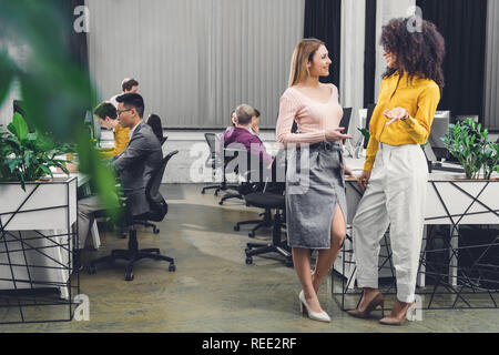 young multiethnic businesswomen talking and smiling each other while colleagues working behind in office Stock Photo