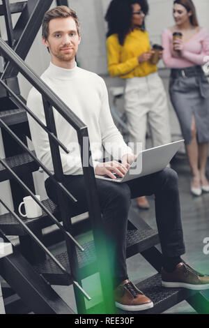 handsome man sitting with laptop on stairs and smiling at camera while female colleagues drinking coffee behind in office Stock Photo