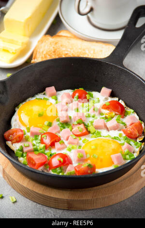 Fried eggs in black pan with ham, cherry tomatoes and spring green onions. Delicious Breakfast with toast and butter. The classic English Stock Photo