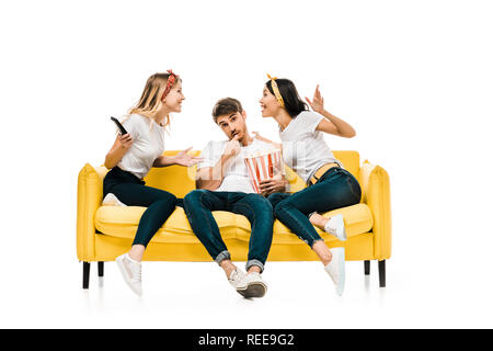 smiling young women talking and man eating popcorn while sitting together on sofa isolated on white Stock Photo