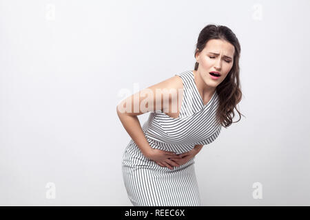 Stomach pain. Portrait of sick unhappy young brunette woman with makeup and striped dress standing with stomach pain and holding her painful belly. in Stock Photo