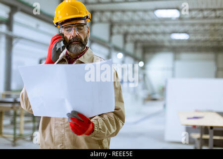 Handsome mature engineer looking at plan and using mobile phone in the factory Stock Photo