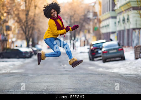 Cheerful young black woman in scarf and sweater is jumping in the middle of the street.