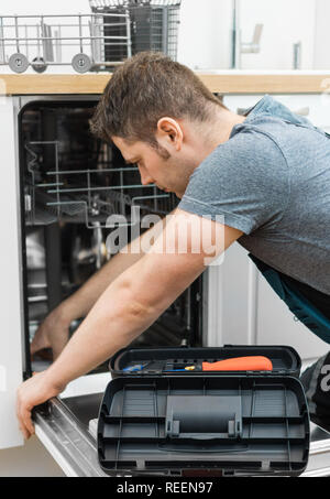 Professional handyman in overalls repairing domestic dishwasher in the kitchen. Stock Photo