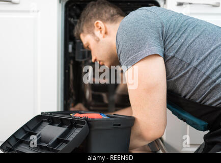 Professional handyman in overalls repairing domestic dishwasher in the kitchen. Stock Photo