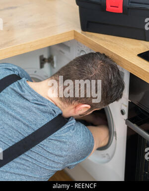 Professional handyman in overalls repairing washing machine in the kitchen. Stock Photo