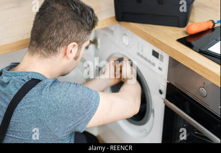 Professional handyman in overalls repairing washing machine in the kitchen. Stock Photo