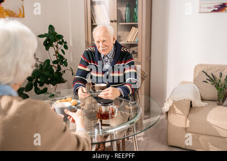 Portrait of cute elderly couple drinking afternoon tea at glass table in living room and enjoying conversation, copy space Stock Photo