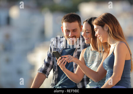 Three happy friends using a smart phone in a town at sunset Stock Photo