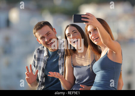Three happy friends taking selfies with a smart phone outdoors in a town outskirts at sunset Stock Photo