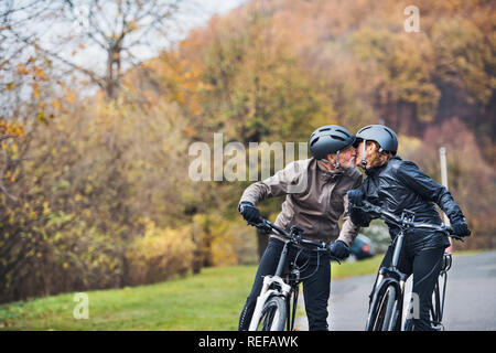 Active senior couple with electrobikes standing outdoors on a road in nature, kissing. Stock Photo