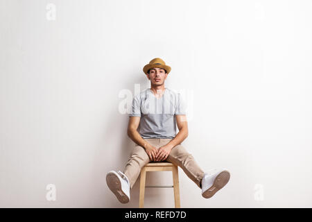 A confident hispanic young man with hat sitting on a stool in a studio. Stock Photo