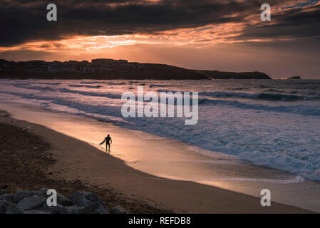 A spectacular sunset as a lone surfer carrying his surfboard walking along the shoreline of Fistral Beach in Newquay Cornwall. Stock Photo