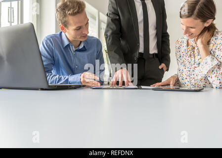 Team of three business people working together reviewing documents, checking their data on laptop and digital tablet while at their white office desk. Stock Photo