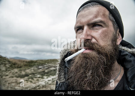 Man with long beard and mustache smoking cigaret. Brutality concept. Hipster on strict face with beard looks brutally while hiking and smoking. Man with brutal bearded appearance, untidy and unshaven Stock Photo