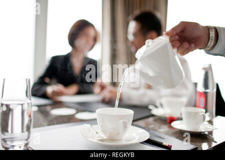 Businessman and woman talking at a conference table, with tea being poured in the foreground. Stock Photo