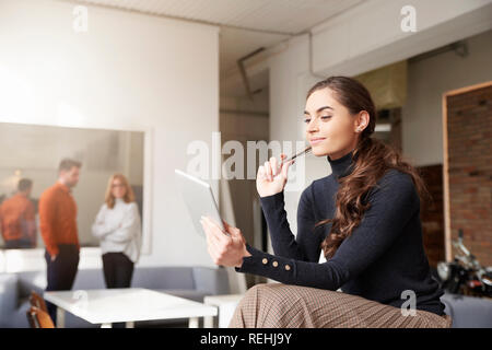Thinking young sales woman holding digital tablet in her hand while sitting at office desk and browsing the internet. Stock Photo