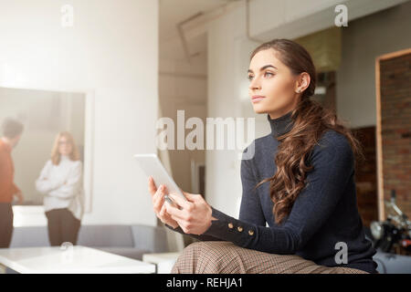 Thinking young sales woman holding digital tablet in her hand while sitting at office desk and browsing the internet. Stock Photo