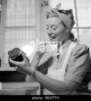 1940s practical tips. A young woman at home looks happy when licking the glue side of a label to put on the glass jar of the jam she has made herself. She is in the kitchen dressed in an apron and has her hair set up in a typical way. The year is 1940 and the world war II is going on. Food and other necessities are rationed, therefore an own supply of food was neccesary.  Photo Kristoffersson 158-1 Stock Photo